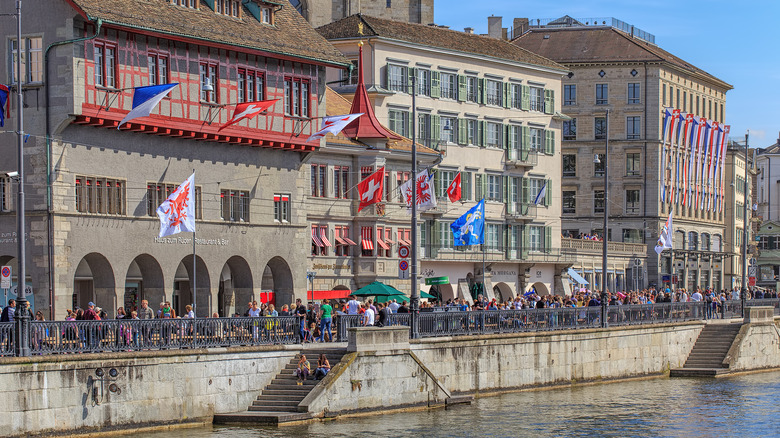 People crowd the street near a river lined with historic buildings in Zurich