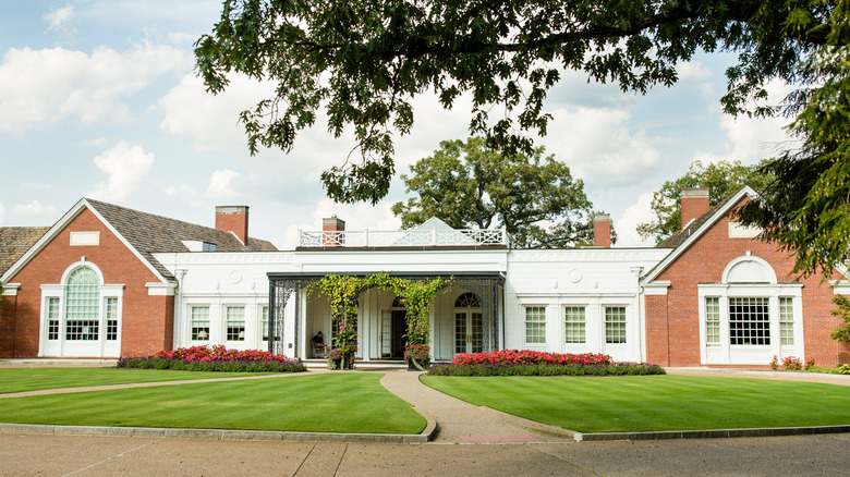 Fox Chapel Country Club, a long building of white and red brick walls behind a lawn with red flowers under a cloudy blue sky