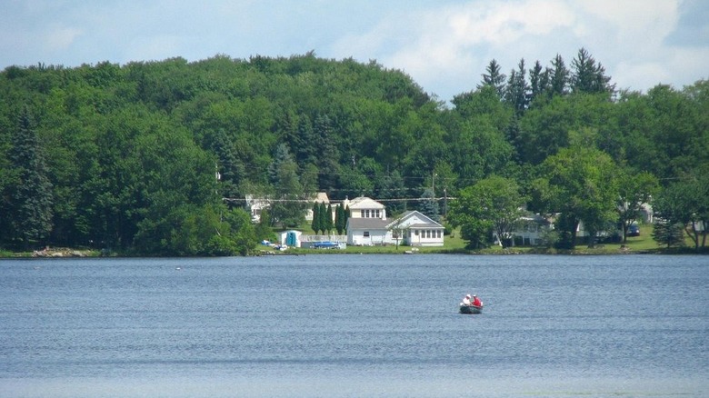 Blue lake with a fishing boat that has two people inside, with homes on the far shore surrounded by green trees under a cloudy blue sky