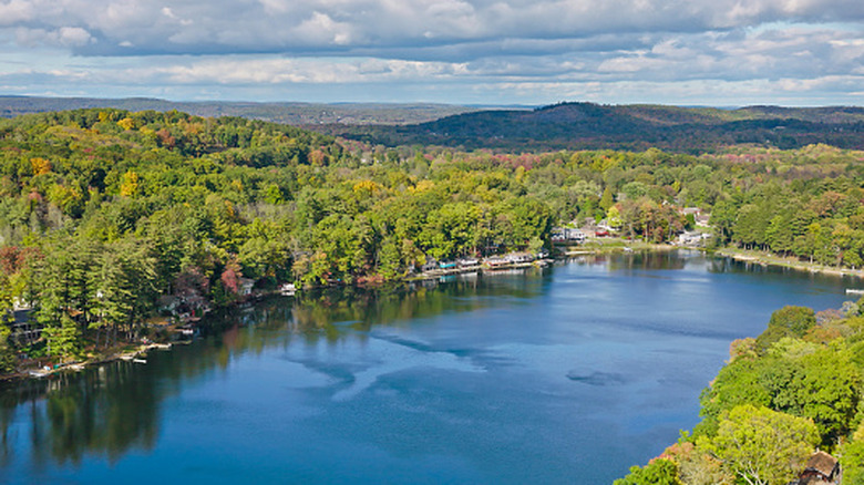 Lake in the Pocono Mountains, with homes along the shore surrounded by trees and green hills in the background, under a cloudy blue sky
