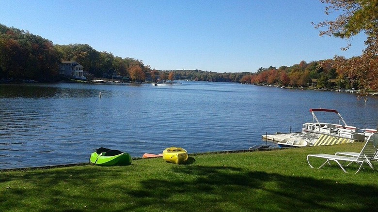 Blue lake under a blue sky surrounded by fall foliage, with grass, a boat, and yellow and green kayaks in the foreground and homes and boats along the shore