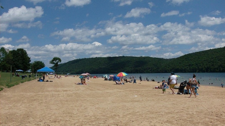 Beach with people, a blue canvas tent, and a rainbow umbrella along the side of a blue lake, with green hills in the background under a cloudy blue sky