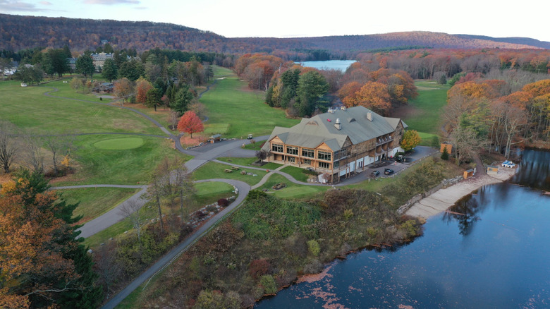Skytop lodge, a large gray-roofed stone-walled building surrounded by lawns and autumn trees, with a lake and beach in the foreground and a lake in front of rolling hills in the background