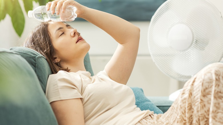 Young woman sitting on a couch holding a cold bottle on her head with fan in the background