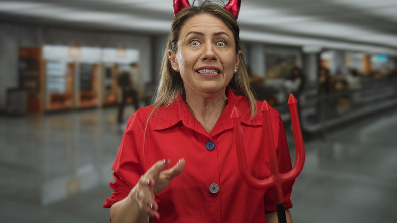 A woman holds a red trident and wears devil horns in a bustling airport terminal