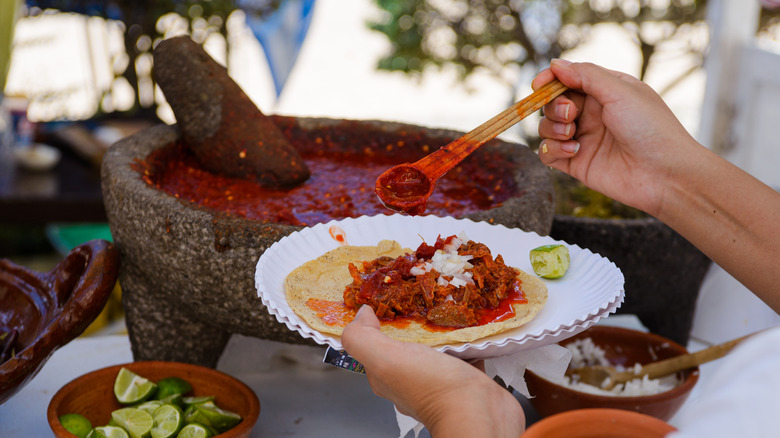 A hand adds spicy red sauce to a delicious-looking taco on a paper plate.