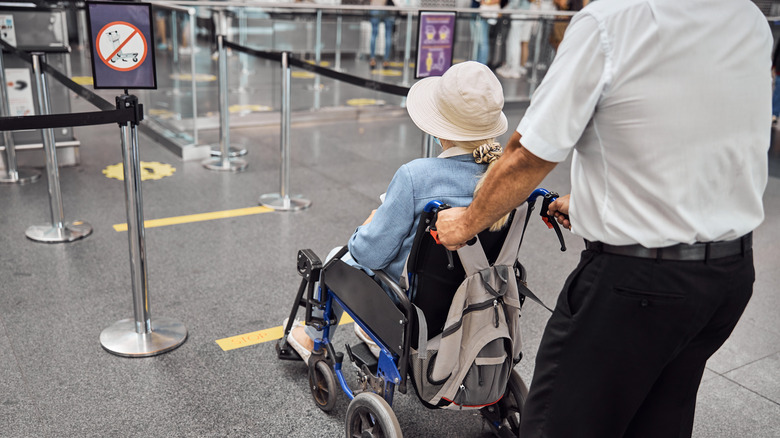 An elderly woman wearing a hat being pushed through the airport on a wheelchair by a uniformed attendant.