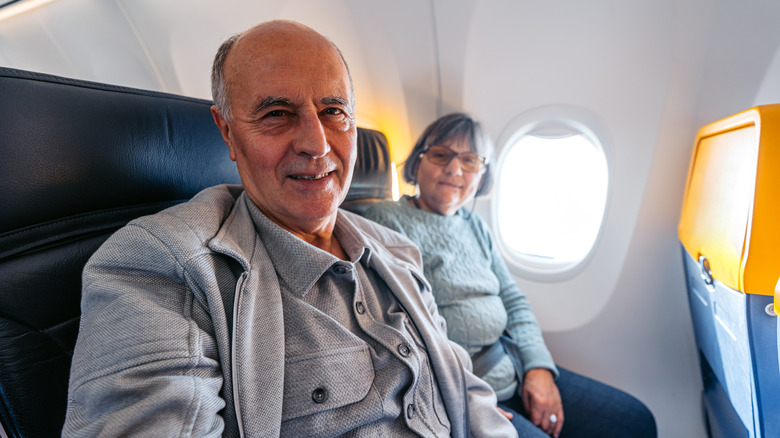A man and woman with grey hair sitting on a plane smiling into the camera.