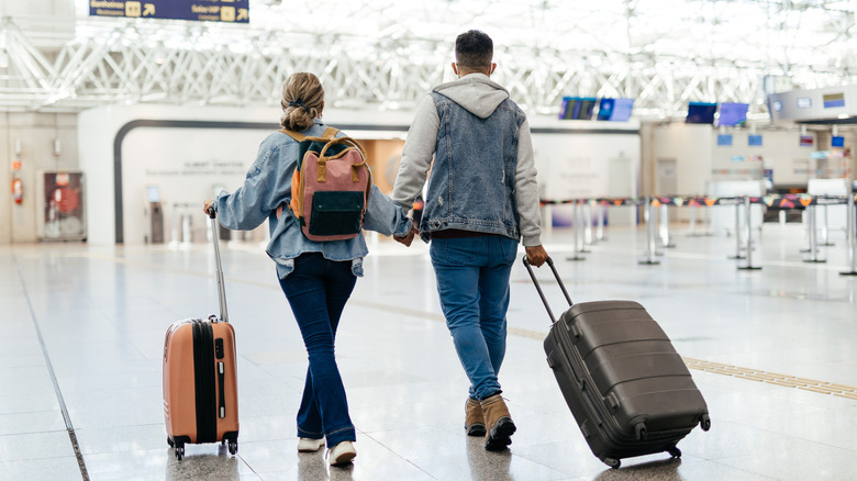 Back view of a Caucasian couple holding hands, pulling luggage, while walking through an airport