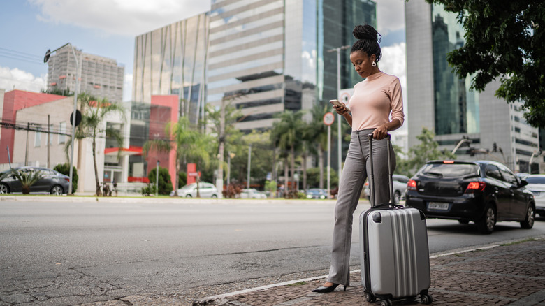 a woman stands on the sidewalk with luggage looking at her phone