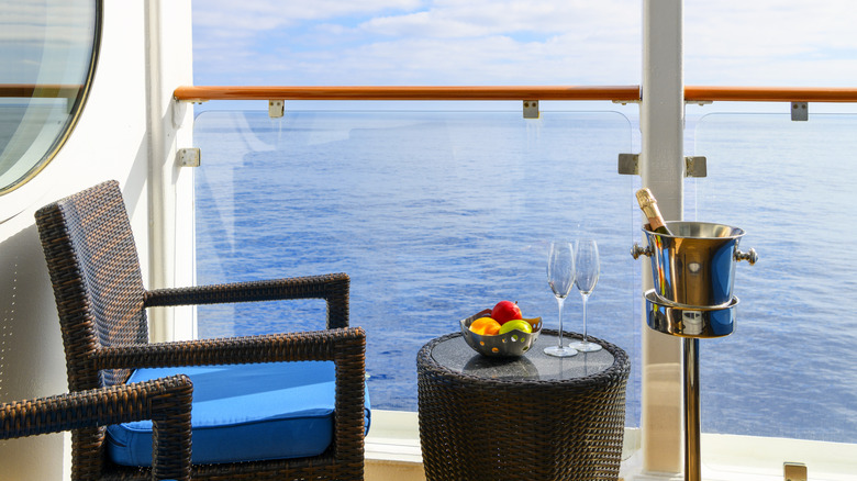 A bowl of fresh fruit, a bottle of champagne in an ice bucket, and two champagne glasses on an oceanfront balcony veranda on a sunny cruise ship.