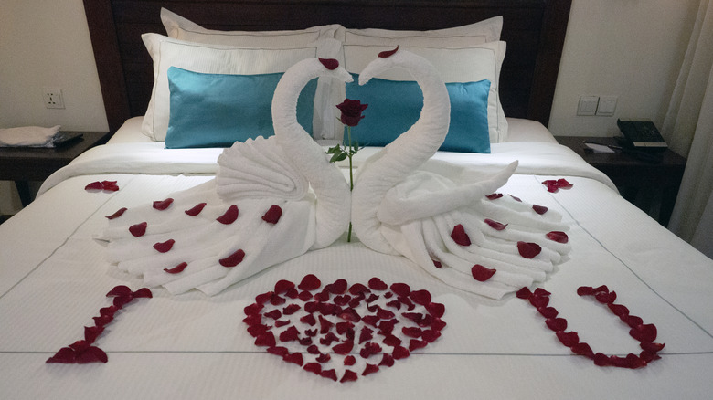 Two towel swans kissing each other on the bed, their heads form a heart. Rose pedals all around.