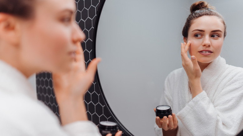 A young woman in a bathrobe looks at herself in the mirror while applying cream to her face