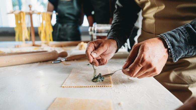 People in pasta-making class surrounding counter with homemade pasta while person places cream spinach on ravioli sheet