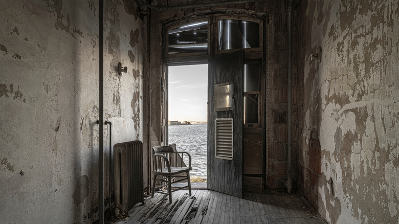 Abandoned hospital room with cement walls and a chair in front of an open door in Ellis Island Immigrant Hospital, New York