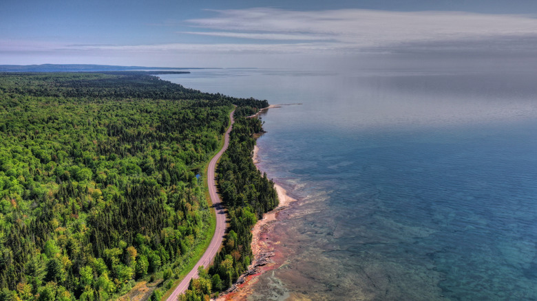 An aerial photo above the Keweenaw Peninsula with thick green forests meeting the vast Lake Superior with a thin beach on the shore and a two-lane road snaking along the shoreline