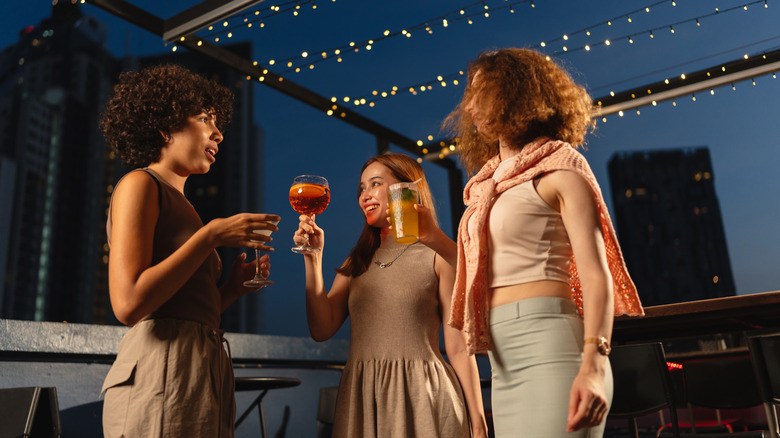 Three women enjoy cocktails and conversation at a rooftop bar during the evening.