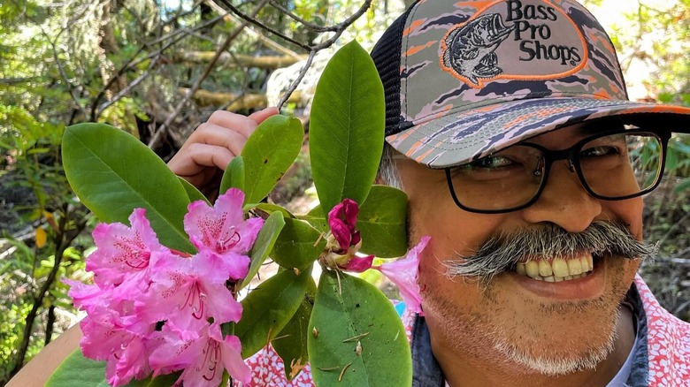 A smiling man holds a rhododendron bloom near his face at Kruse Rhododendron State Natural Reserve, in California.