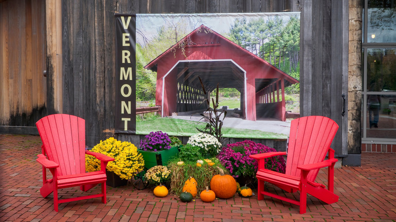 Red Adirondack chairs, wildflowers, and pumpkins stand in front of a banner of a covered bridge in Vermont