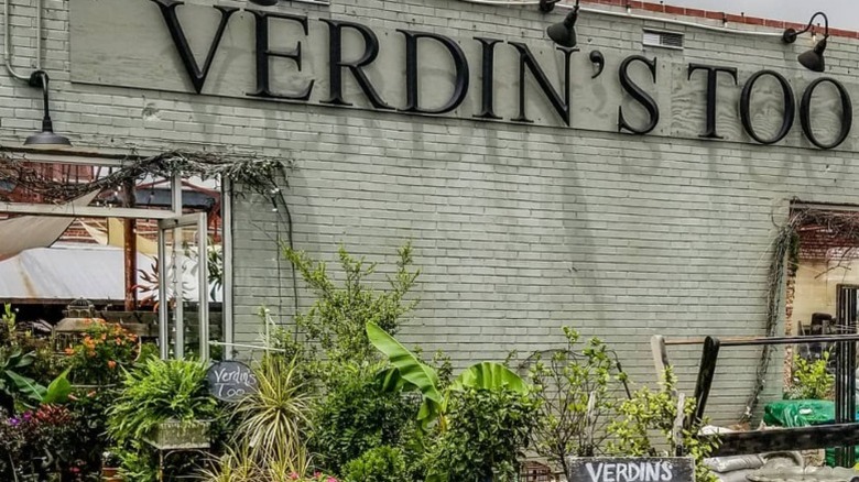 Entrance to Verdin's Too, with a sign on a brick wall and green plants.