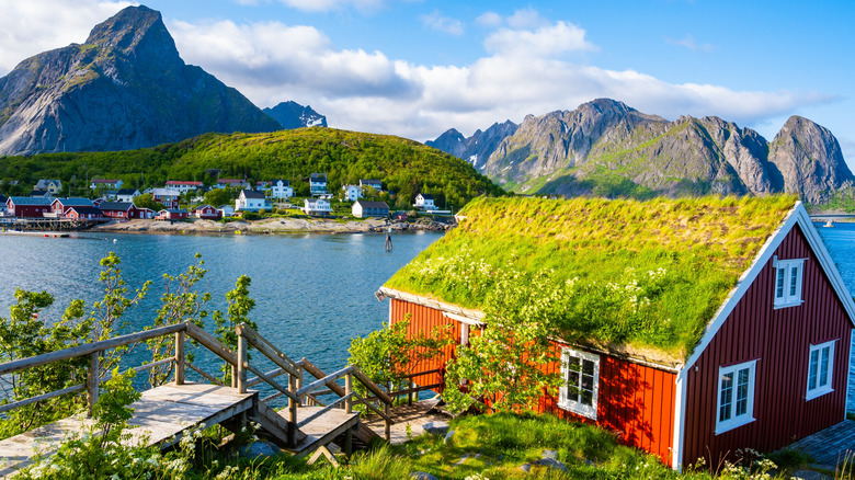 Red cabin with grass roof overlooking a blue lake with houses across the shore, surrounded by lush green hills and towering mountains, Norway