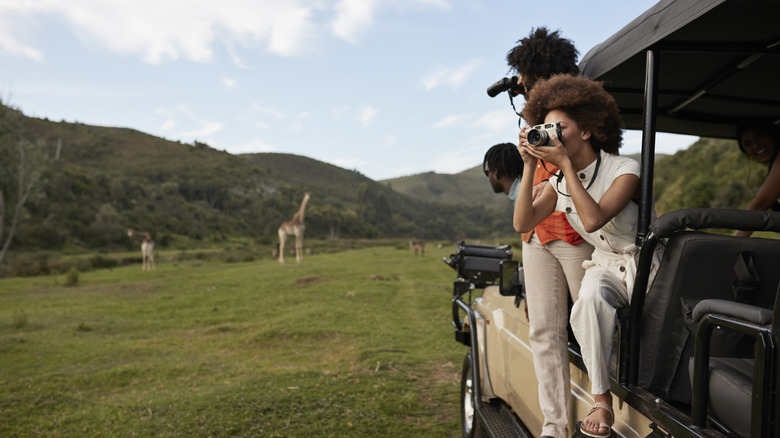 Young woman photographing with camera while man looking through binoculars from safari vehicle in wildlife reserve.