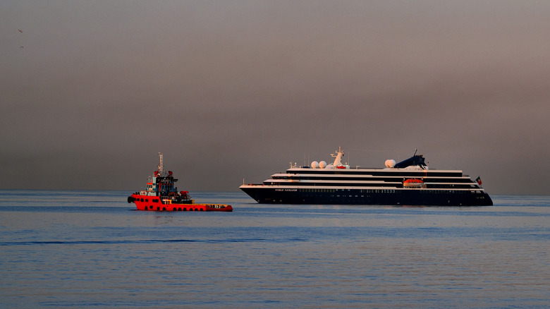 An Atlas Ocean Voyages cruise ship arriving at the port of Piraeus, cruising next to a tugboat on calm water.