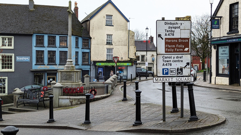 A sign gives directions in English and Welsh on a street corner without houses and a statue in the background in Narberth, Wales