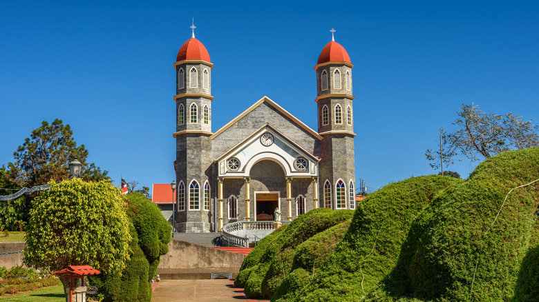 a gray stone church with red domes on two towers, surrounded by a topiary landscape