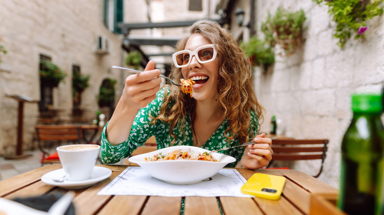 A woman in thick-framed sunglasses sits a wood table and gleefully eats pasta from an elegant soup-plate