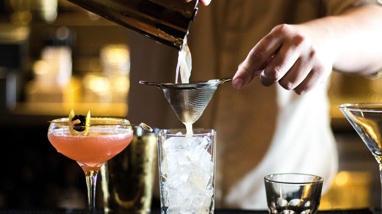 Mixologist pouring drinks in a tall glass with ice, next to other cocktail and shot glasses with a blurred bar setting in the background.