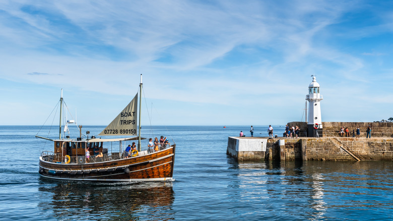 People on a wooden boat drifting past a white lighthouse and harbor wall in Mevagissey, Cornwall