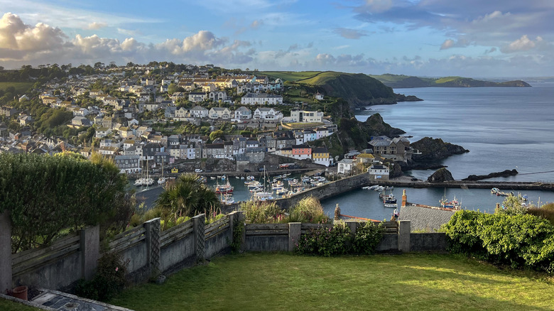 White houses on a hillside overlooking a harbor, and sea wall in Mevagissey in Cornwall, with hills in the distance