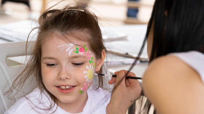 A young girl getting her face painted by a woman at a festival.