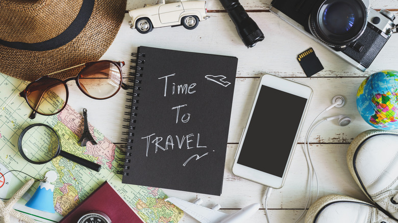 Overhead image of various travel accessories, including a sunhat, sunglasses, map, camera, SD card, smartphone, flashlight, and headphones. In the center is a black notebook labeled 