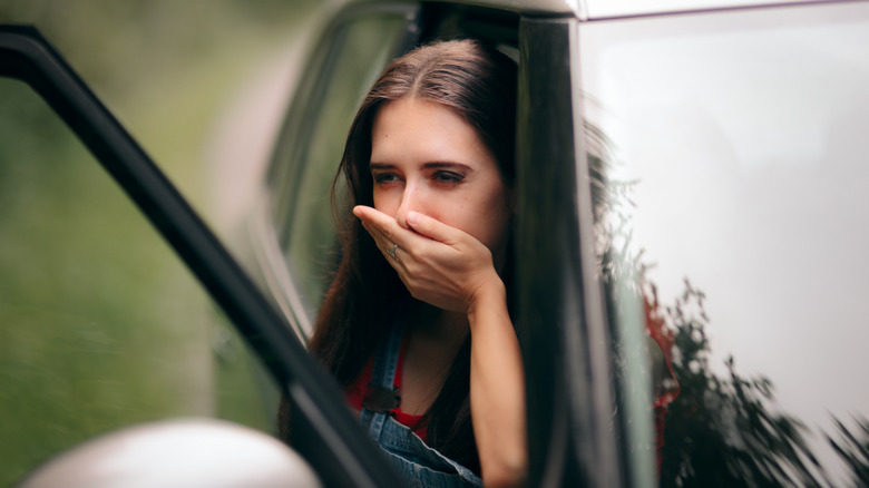 Woman sitting in a car with the door open, leaning out of the window while covering her mouth with her hand, appearing to feel nauseous or carsick