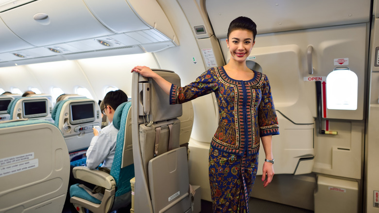 Singapore Airlines crew member smiling inside A380 cabin next to closed aircraft door while passengers sit in seats