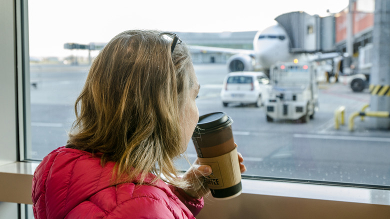 A woman drinking a takeaway coffee while looking through a window at a plane at an airport.