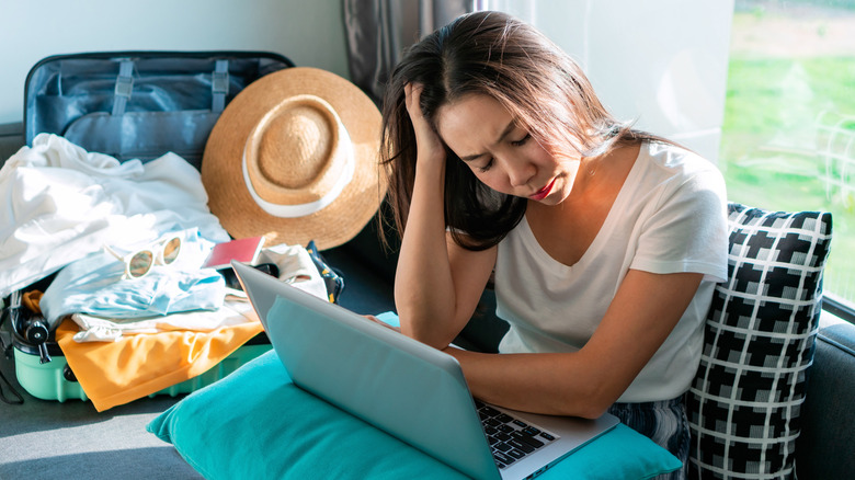 A young woman looks stressed as she peers at her laptop, with an overflowing packed suitcase visible behind her
