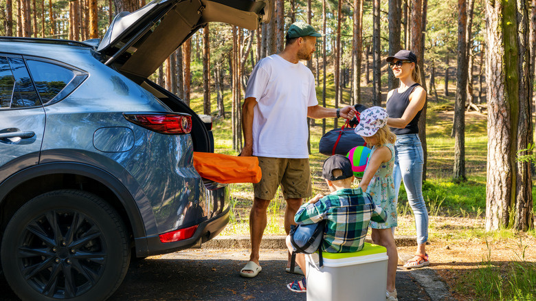 Man, woman, and two children all wearing hats packing the trunk of a car parked by trees