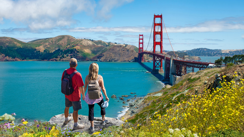 A hiker couple stands holding hands while looking toward the Golden Gate Bridge from a lookout at Lands End