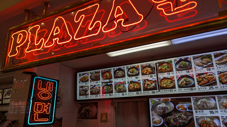 A food stall at the Koreatown Plaza food court with a neon sign and menu board with photos
