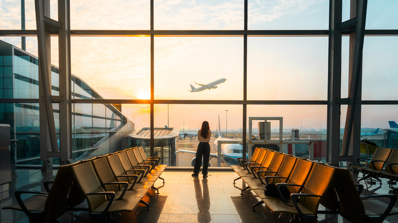 A woman looking out an airport window and watching a plane take off
