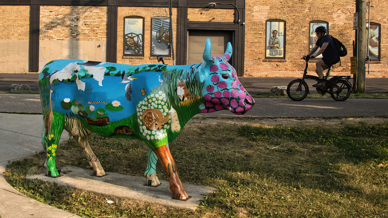 A colorfully decorated cow sculpture stands in the foreground of a Wisconsin street scene as a cyclist pedals in the background