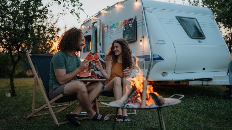 Couple eating in front of campsite fire with an RV in the background