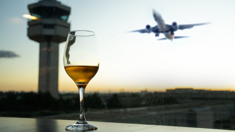 A close-up of a glass of rose wine sitting on a counter in front of a large window at an unidentified airport, with an airplane taking off and an air traffic control tower visible in the background
