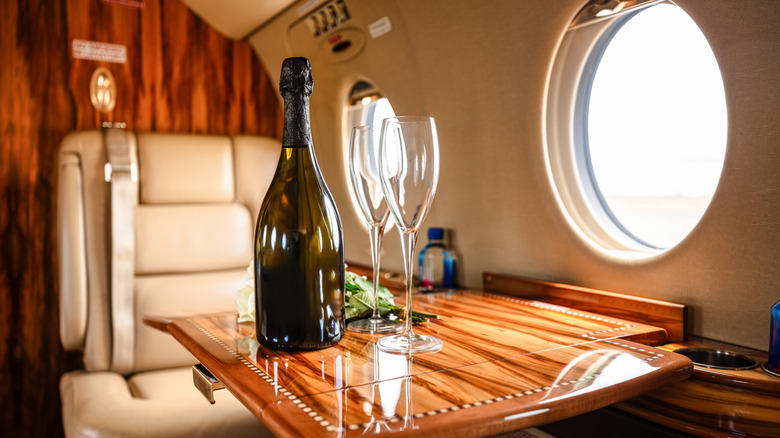 A wide shot of the interior of a private jet, showing leather seats, warm wood accents, and a sleek design, with a bottle of champagne and two glasses set out on the table for passengers