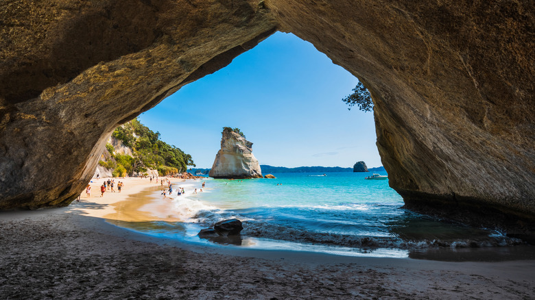 Natural rock arch framing a sandy beach with turquoise waters, sunbathers, and a distant rock formation, Cathedral Cove