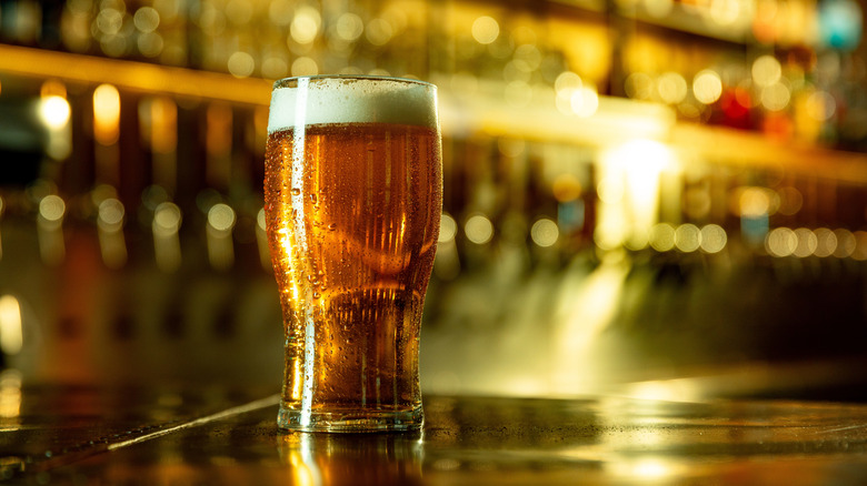 Pint of beer with foam and cold drops standing in front of bar taps and shelves