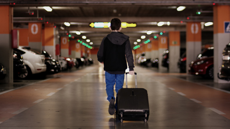 Male traveler with luggage walking toward parked car in a covered airport parking garage.